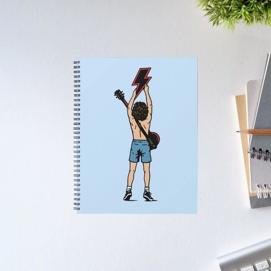 Notebook with a cover featuring an illustration of a person holding a guitar on a light blue background, placed on a desk. AC/DC Notebook The Banyan Tee