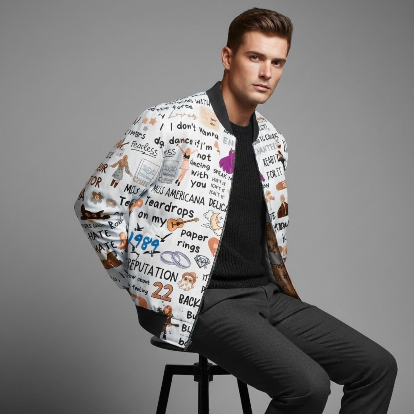 Man wearing a patterned Taylor Swift bomber jacket sitting on a stool against a gray background